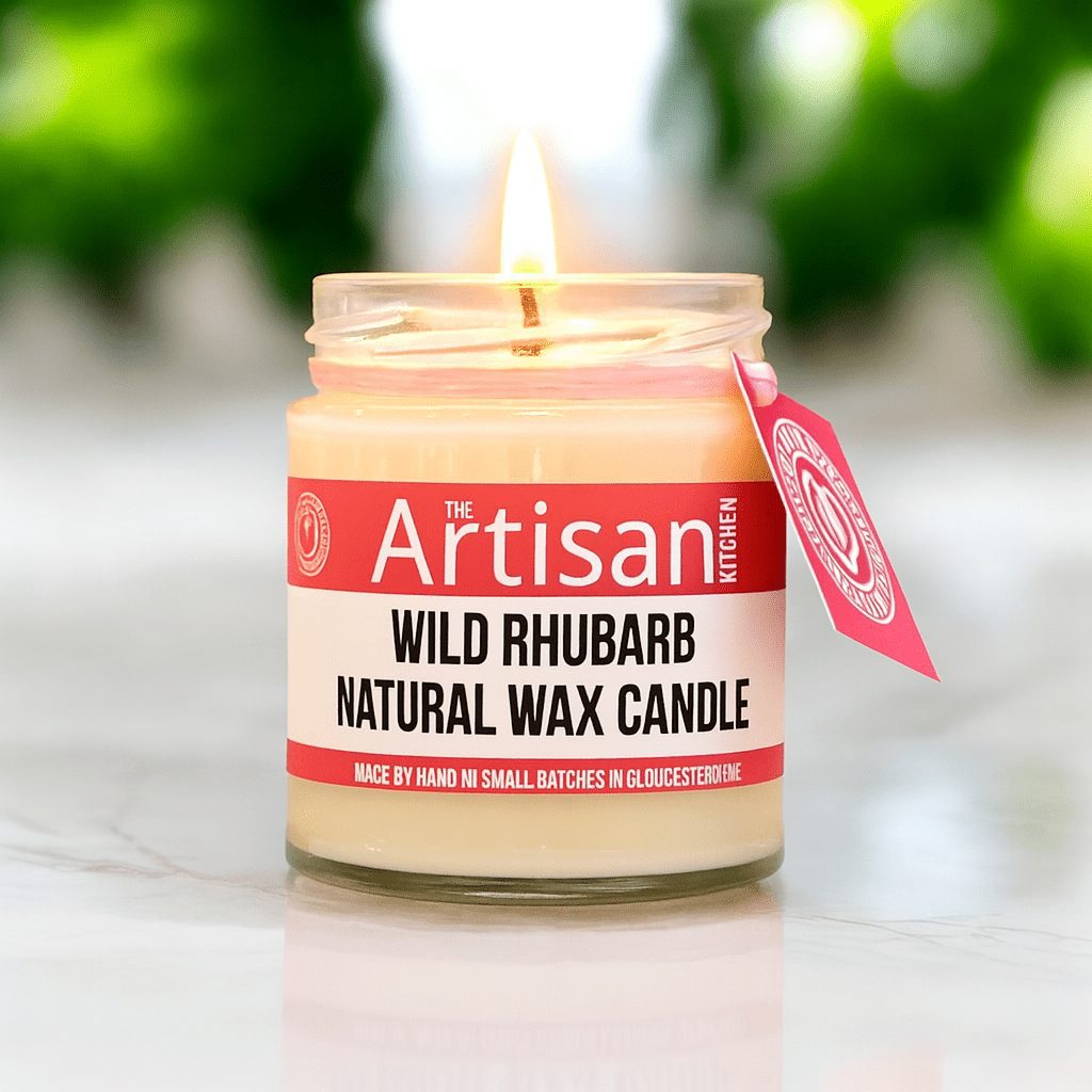 The artisan Kitchen wild rhubarb natural wax candle lit on a white marble counter with blurry greenery in the background