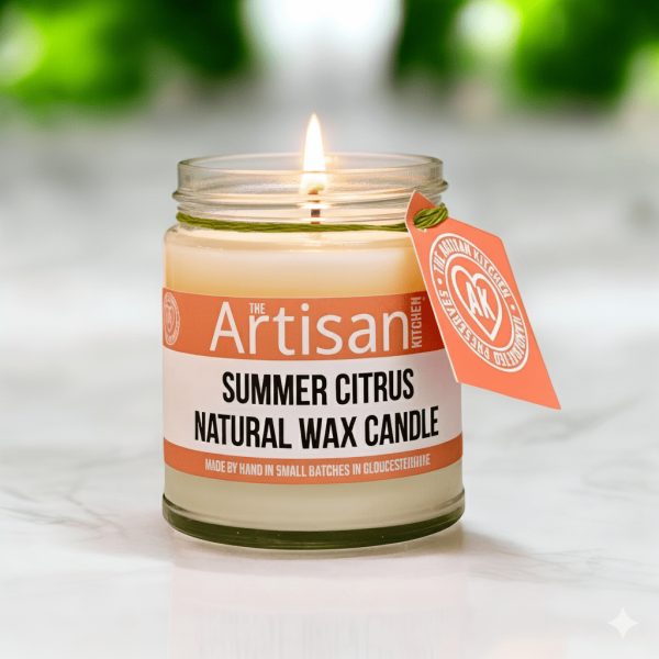 The artisan kitchen summer citrus candle in a glass jar lit sitting on a white marble countertop
