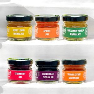 Mini preserves gift box from The Artisan Kitchen featuring six mini jars of honey lemon marmalade, apricot jam, lime lemon vanilla marmalade, strawberry jam, blackcurrant sloe gin jam and summer citrus marmalade, each with Artisan Kitchen branding and award rosette on a white background.